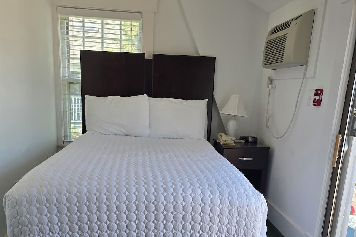 A neatly made bed with a white quilt, a bedside table with a lamp, and a window with blinds.