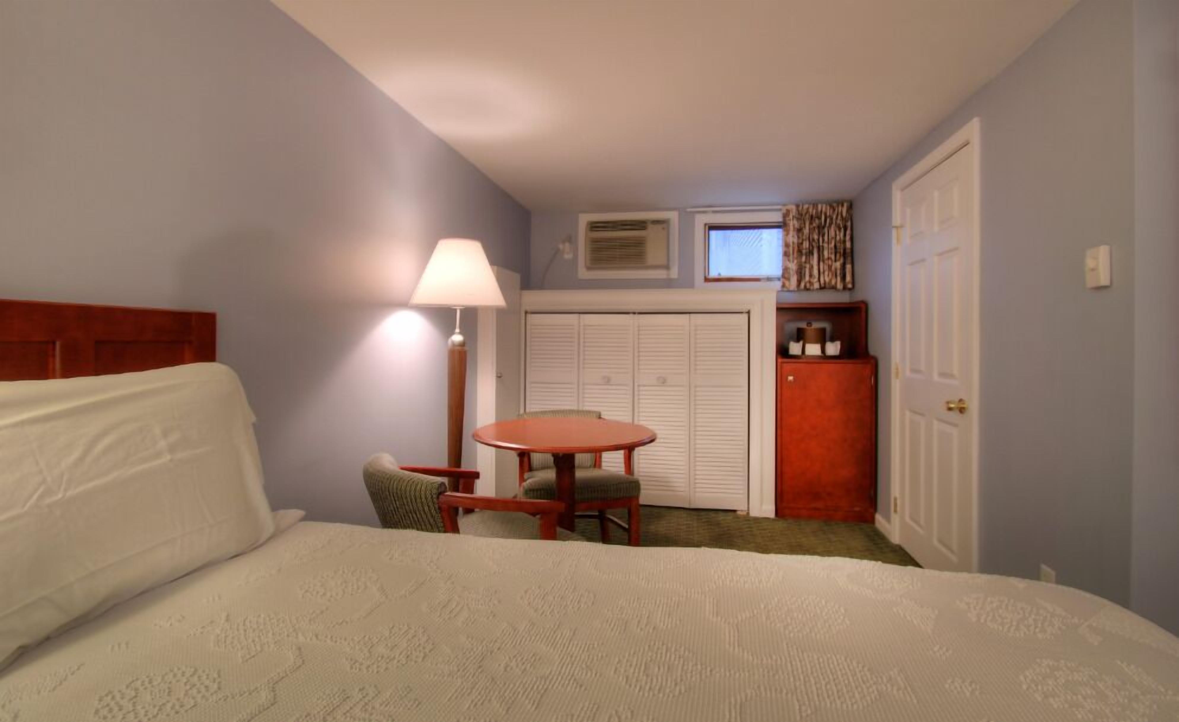 A hotel room with a bed, a desk, a chair, a lamp, a closet, and a window.
