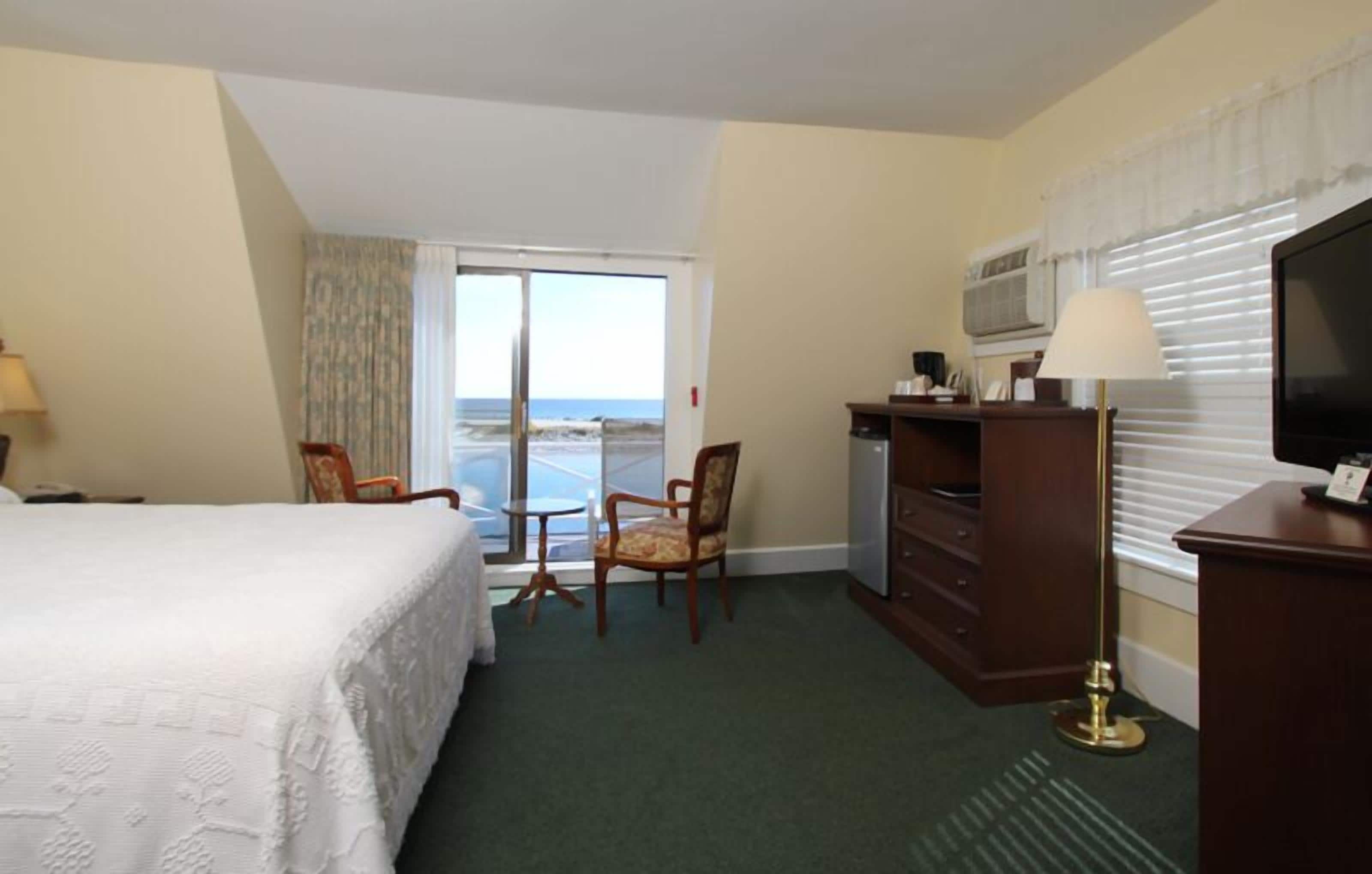 A hotel room with a bed, a desk with a television, a chair, and a view of the ocean through a sliding door.