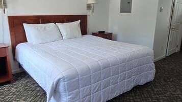 A hotel room with a large bed, a wooden headboard, a nightstand, a lamp, a wall-mounted light, a door, and a patterned carpet.