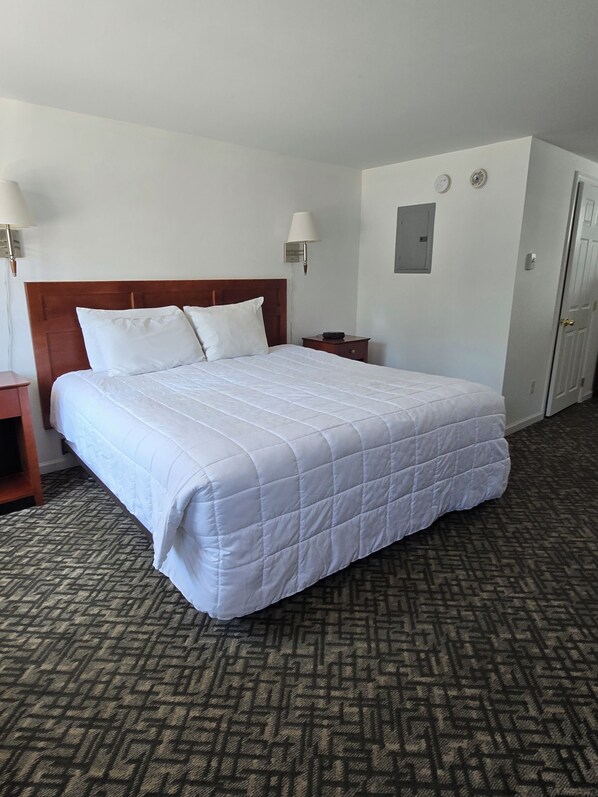 A hotel room with a large bed, a wooden headboard, a nightstand, a lamp, a wall-mounted light, a door, and a patterned carpet. - Terrace by the Sea (Ogunquit)