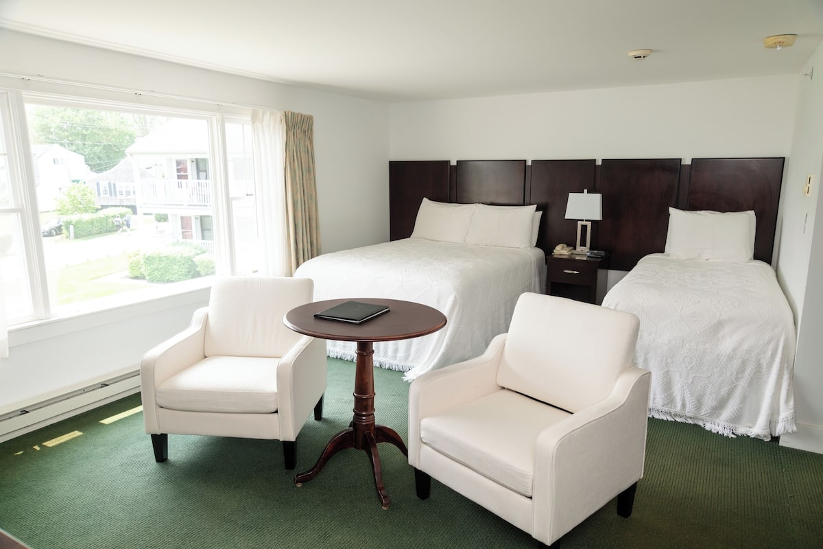 A hotel room with two beds, a small round table, two armchairs, a nightstand with a lamp, and a view of the outside.