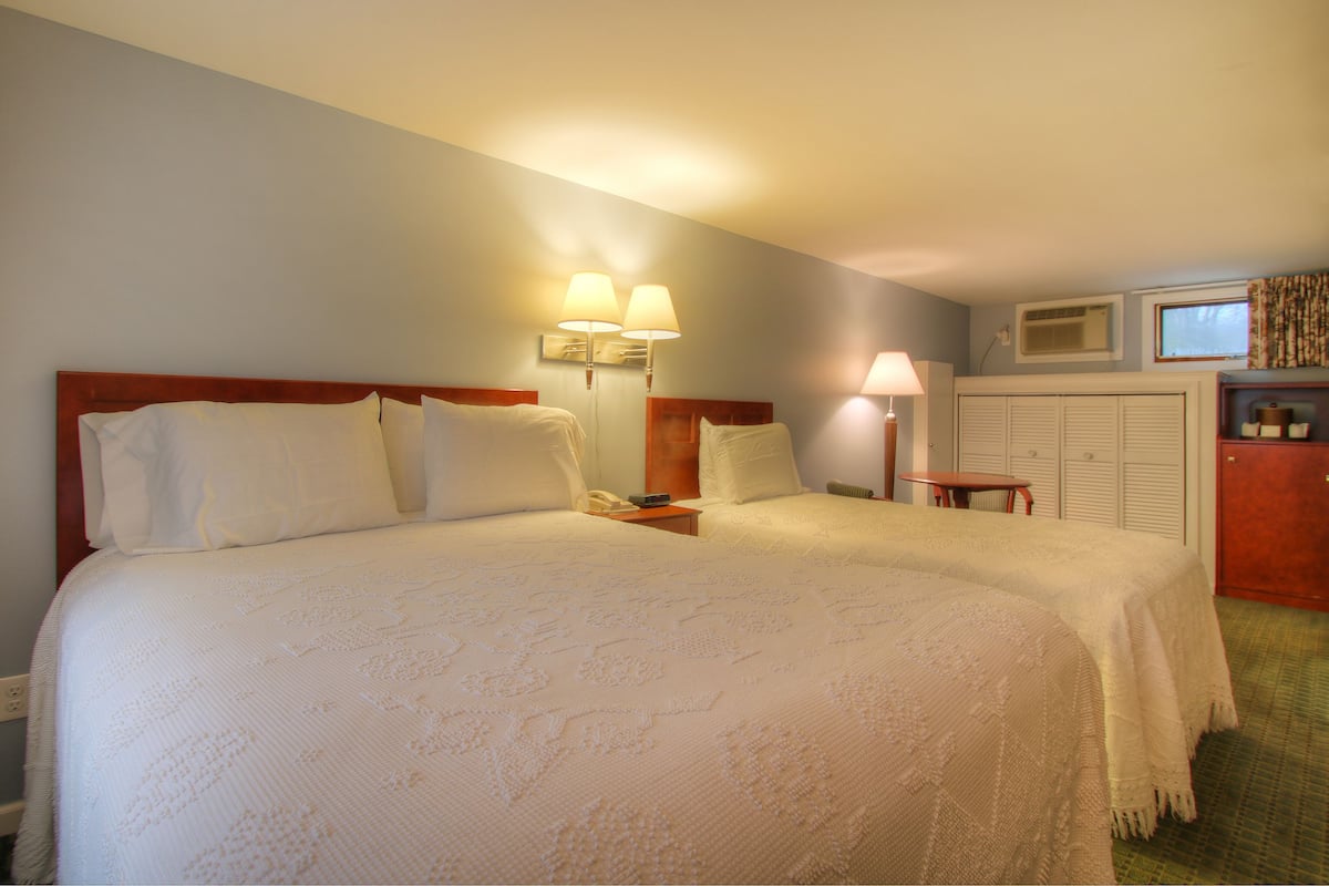 A hotel room with two beds, a small desk, a lamp, a wardrobe, and a window.