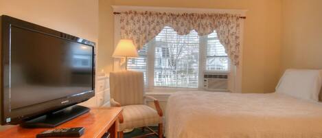 A hotel room with a bed, a TV on a wooden table, a chair, a window with floral curtains, and a lamp.