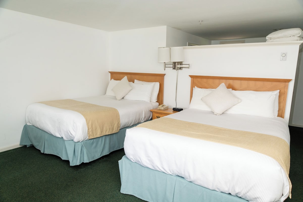 A hotel room with two beds, each with a wooden headboard, white bedding, and a beige throw blanket. There is a nightstand with a phone and a lamp between the beds.