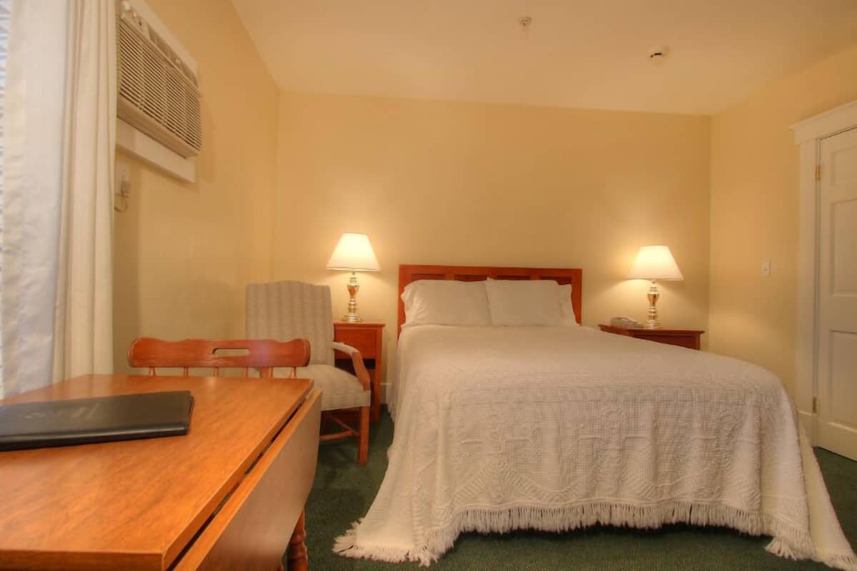 A hotel room with a bed, two bedside lamps, a wooden desk, a chair, and a window with blinds.