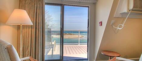 A hotel room with a large sliding glass door leading to a balcony with a view of the beach. There are two chairs and a small table on the balcony.