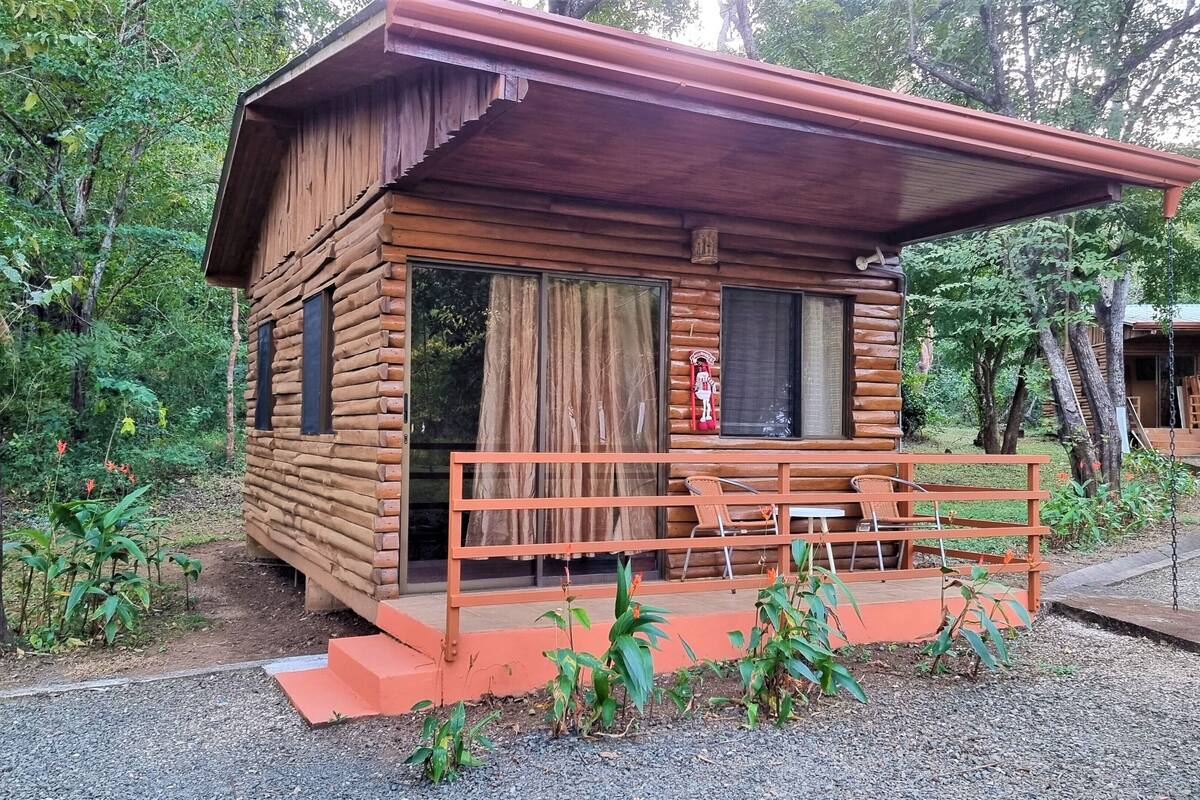 Una cabaña de madera con un porche, rodeada de árboles.
