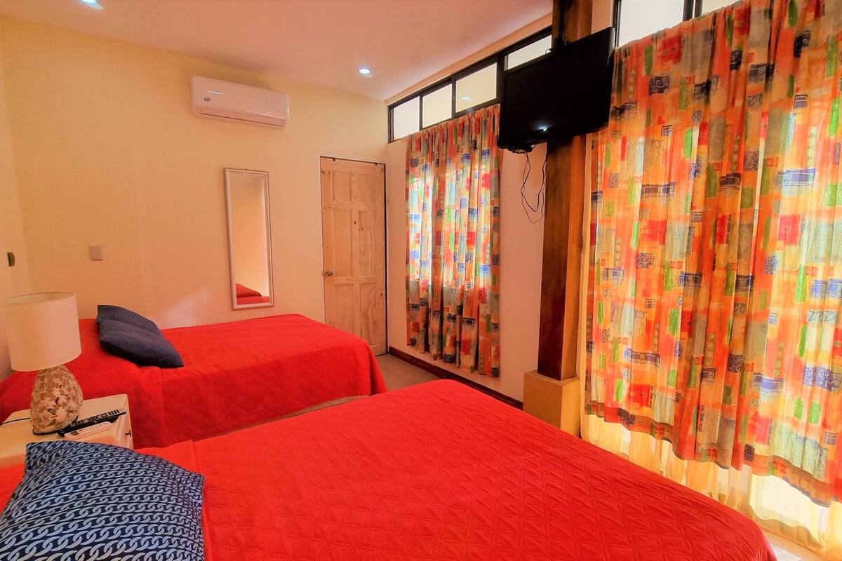 Habitación de hotel con dos camas, televisión, espejo y ventana con cortinas coloridas.