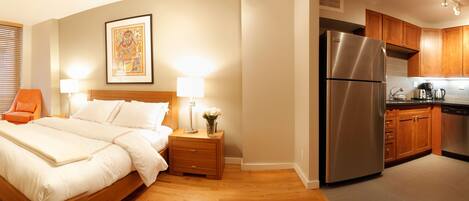 A hotel room with a bed, bedside table, lamp, window with blinds, and a kitchen with a refrigerator, oven, and microwave.