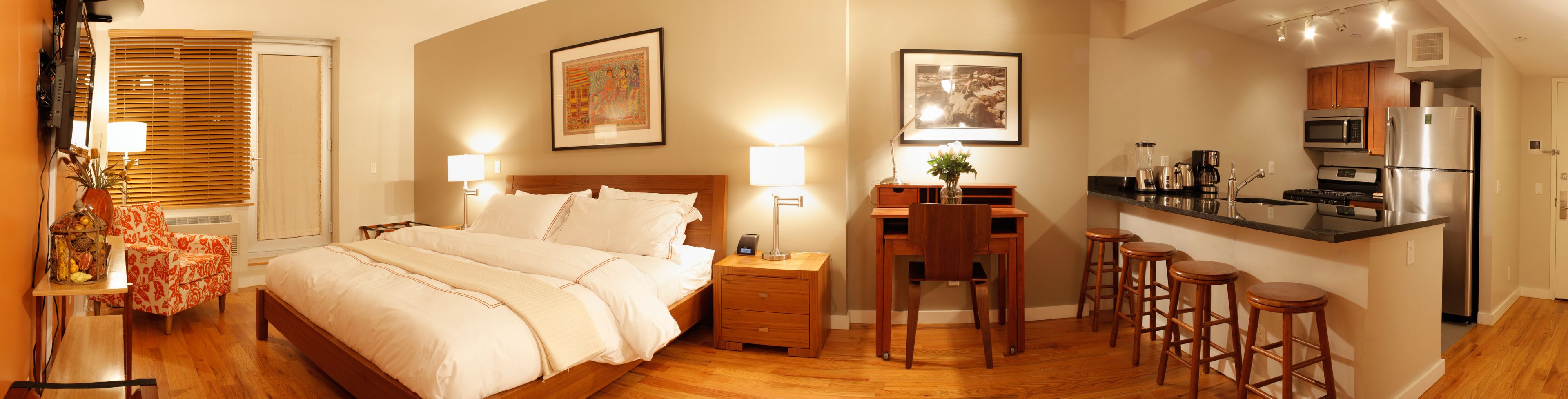 A hotel room with a bed, bedside tables, a desk with a chair, a kitchen area with a refrigerator, microwave, and coffee maker.