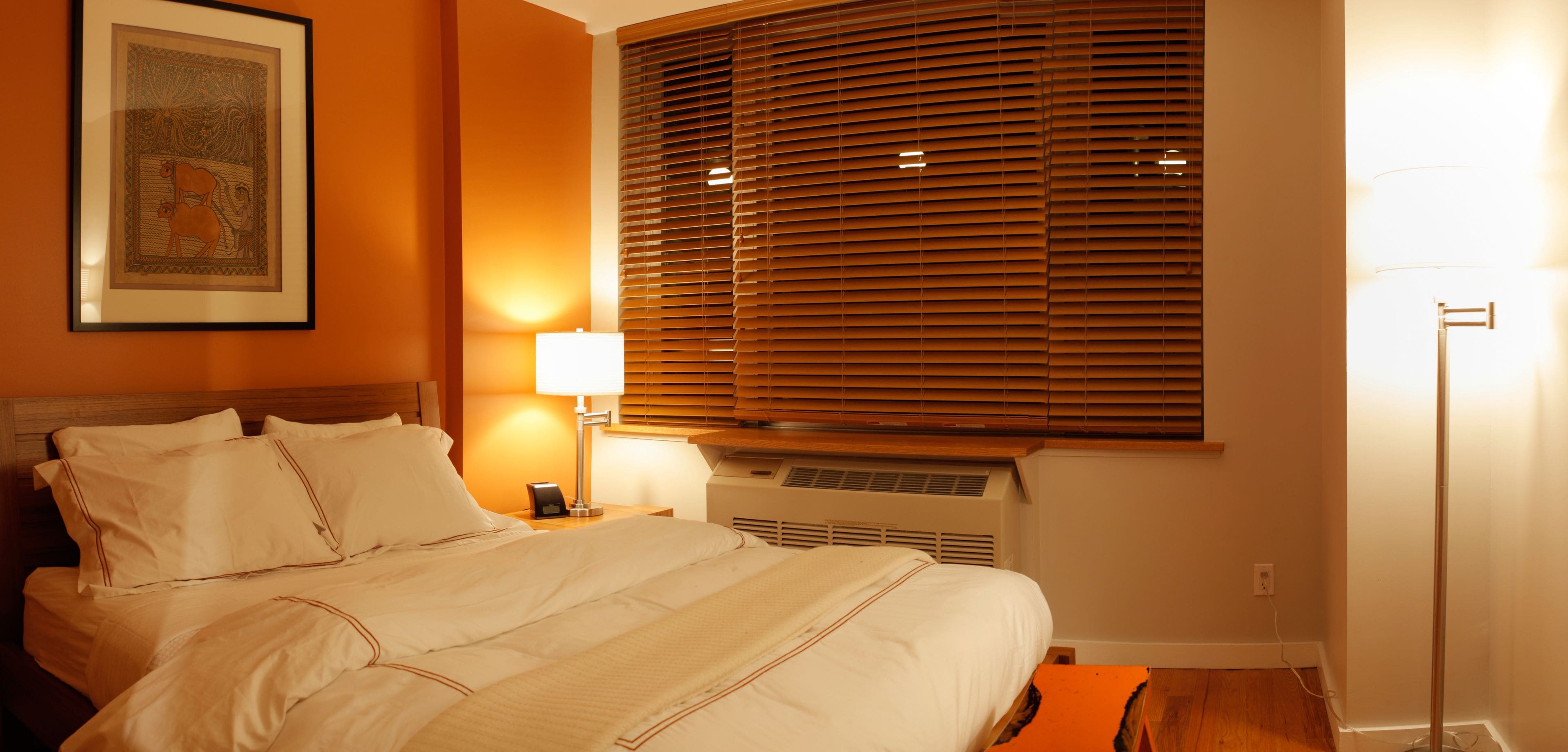 Une chambre d’hôtel comprenant un lit, une table de chevet avec une lampe, une fenêtre avec des stores et un climatiseur fixé au mur.