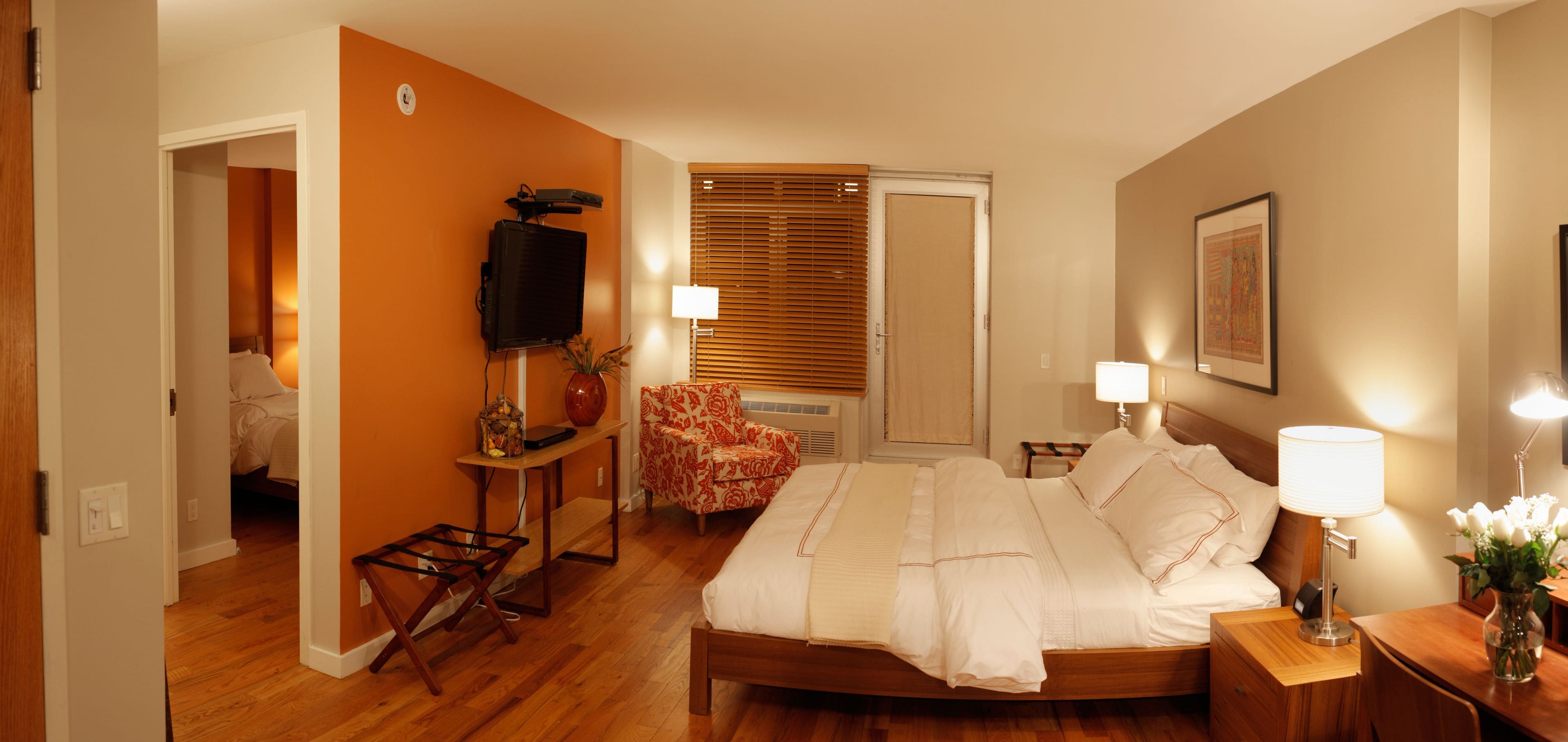 Une chambre d’hôtel avec un lit en bois, un bureau avec une lampe, une chaise, un téléviseur et une fenêtre avec des stores.