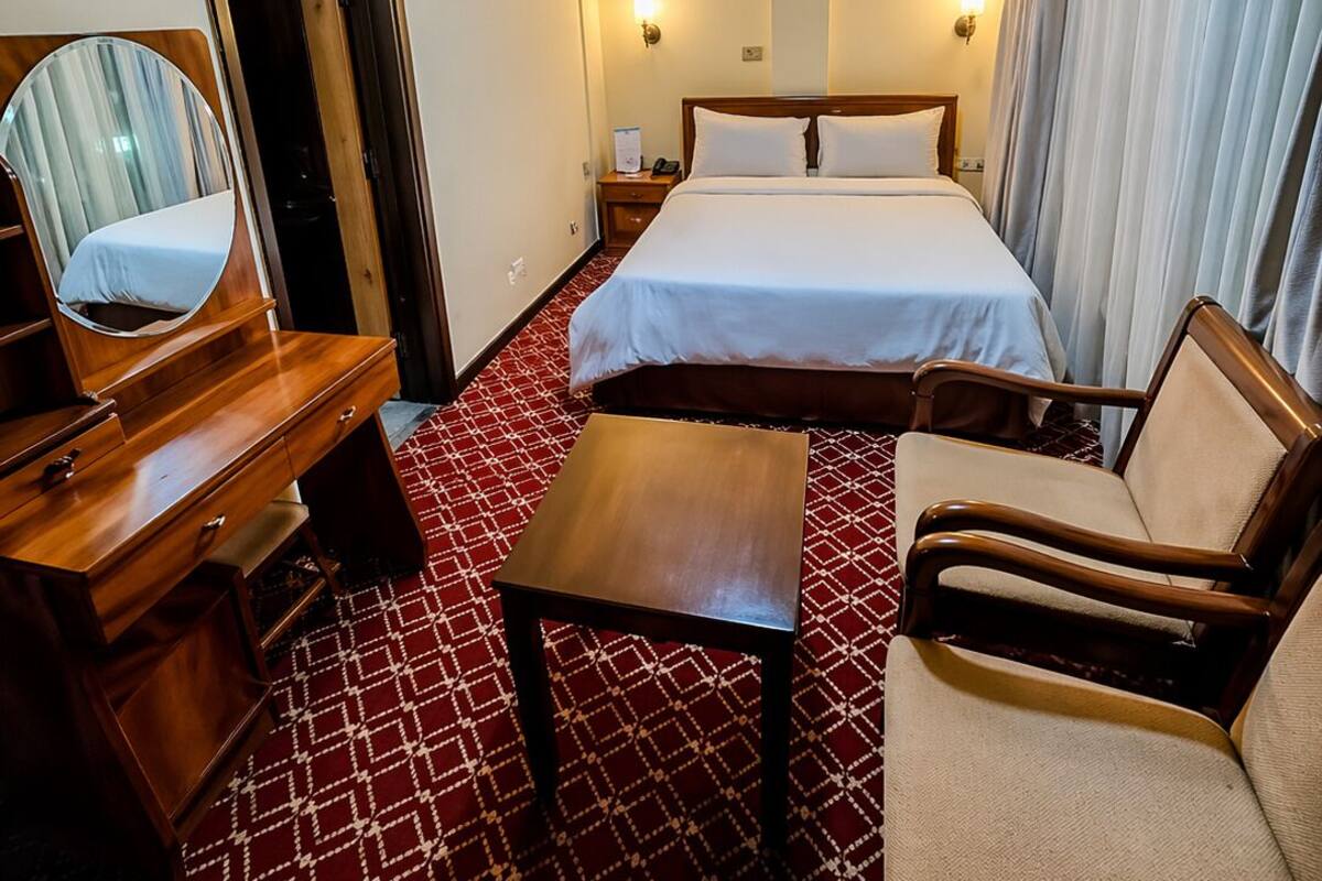 Quarto de hotel com cama, escrivaninha, cadeira, espelho e um tapete vermelho estampado.