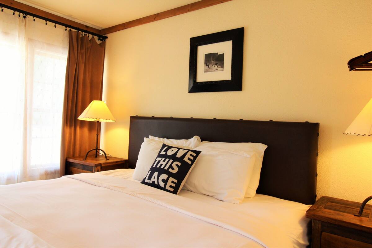 A neatly made bed with white linens, a pillow with the text "LOVE THIS PLACE", a framed picture on the wall, and a lamp on a bedside table.