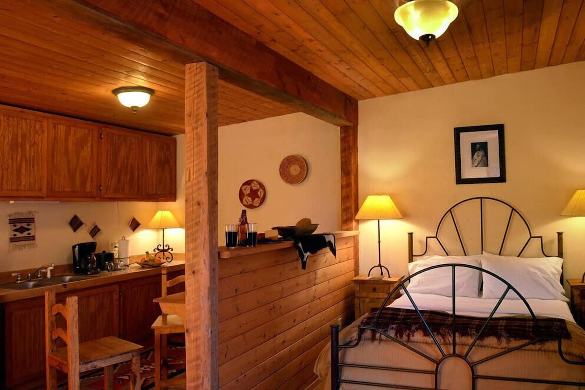 A cozy room with a wooden ceiling, a bed with a metal frame, a nightstand with a lamp, and a kitchen area with wooden cabinets and a sink.