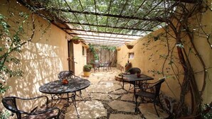 Un patio cubierto con suelo de piedra, sillas y mesas de metal, rodeado de muros con plantas trepadoras.
