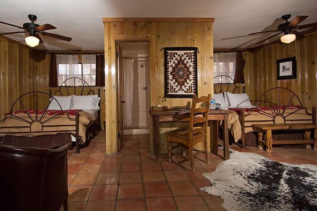 A rustic room with two single beds, a wooden table, chairs, ceiling fans, and a patterned wall hanging.
