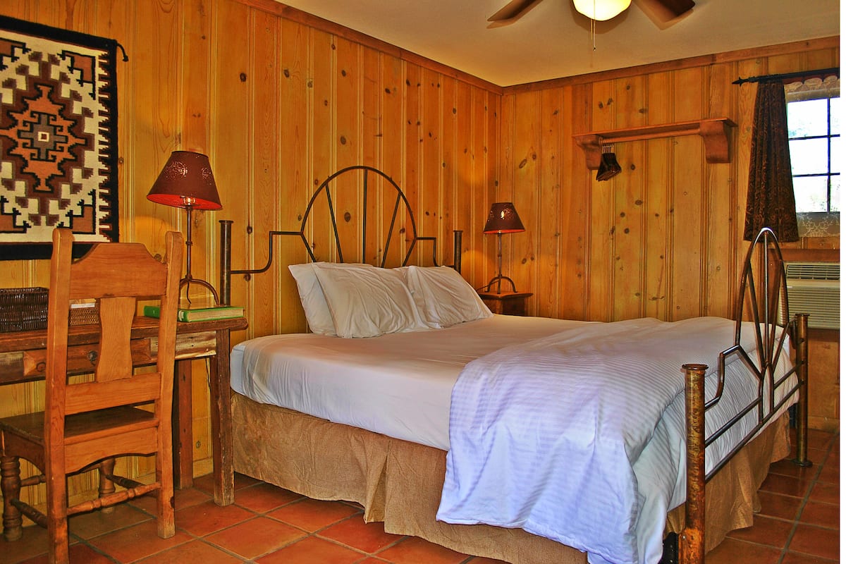 A wooden-paneled room with a bed, two bedside lamps, a chair, and a quilted wall hanging.