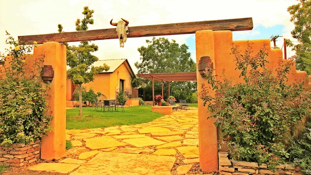 Un patio con un sendero de piedra que lleva a un edificio, un arco de madera con una decoración de calavera y follaje circundante.