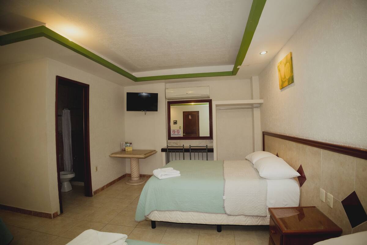 A hotel room with a bed, a bedside table, a TV mounted on the wall, and a bathroom visible through an open door.