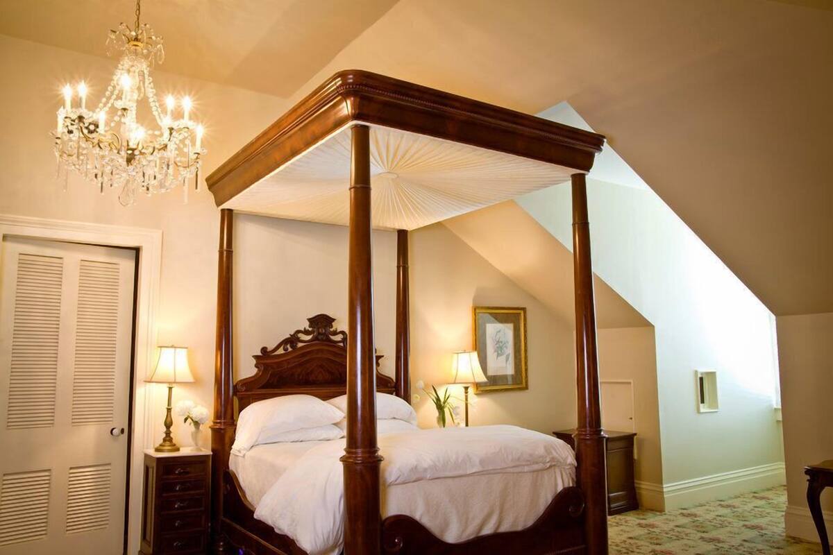 A four-poster bed with a wooden frame and white bedding, a chandelier, and a small table with a lamp.