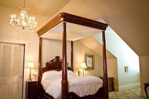 Ein Bett mit Baldachin, dessen Holzbettgestell mit weißer Bettwäsche bezogen ist, eine Kristallleuchter und ein kleiner Tisch mit einer Lampe.