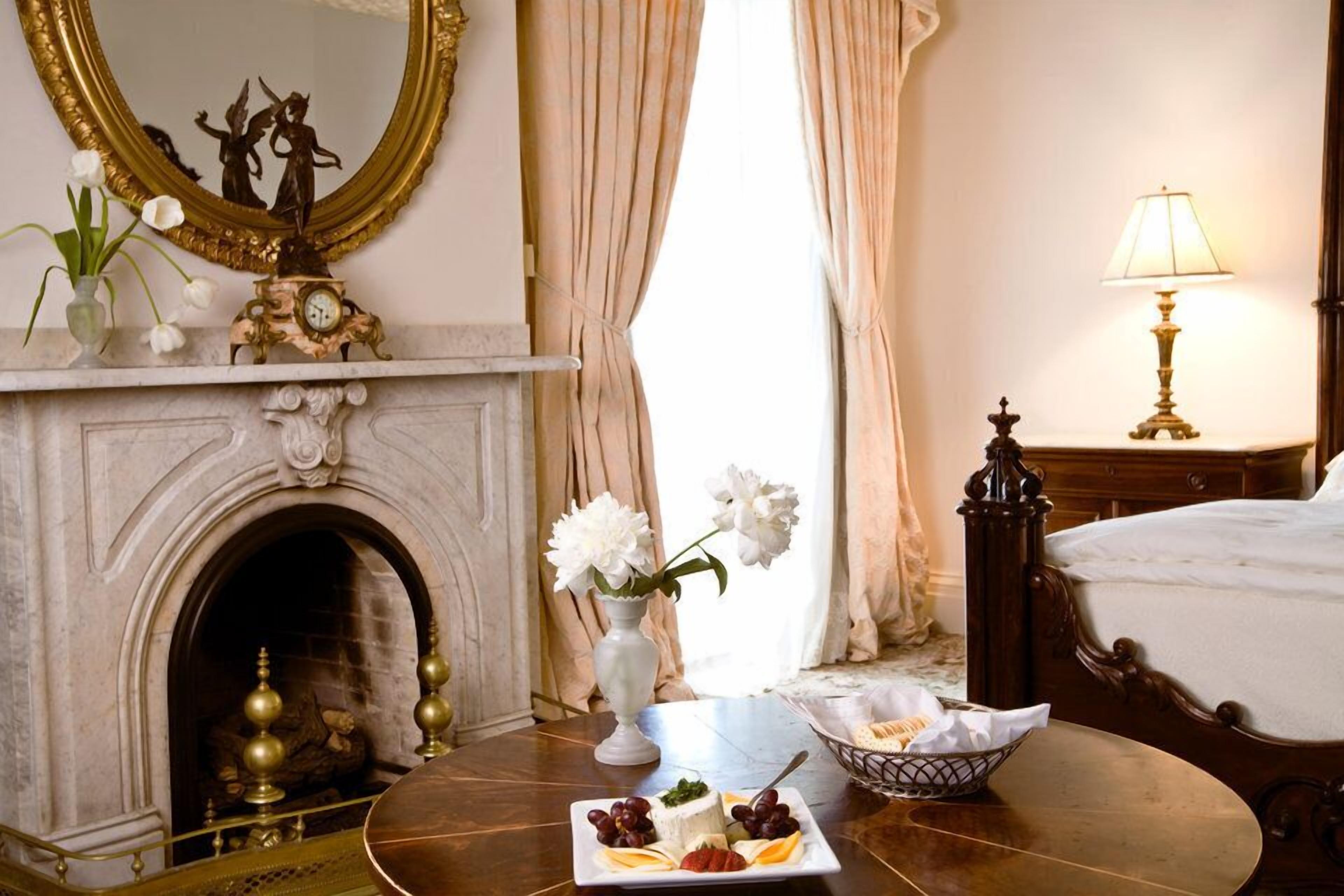 A hotel room with a marble fireplace, a bed with a white bedspread, a bedside table with a lamp, and a dining table with a plate of fruit and a vase of flowers.