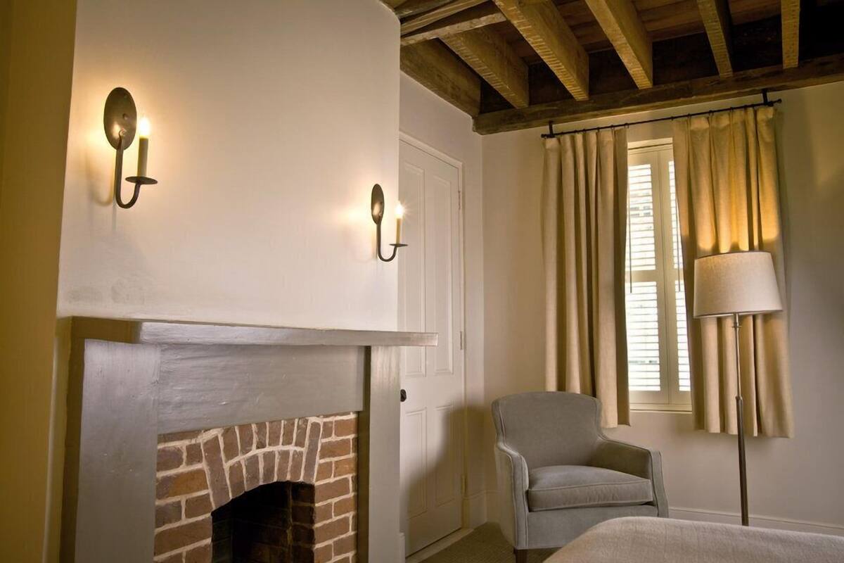 A room with a brick fireplace, two wall-mounted sconces, a grey armchair, a bedside table, and a floor lamp.