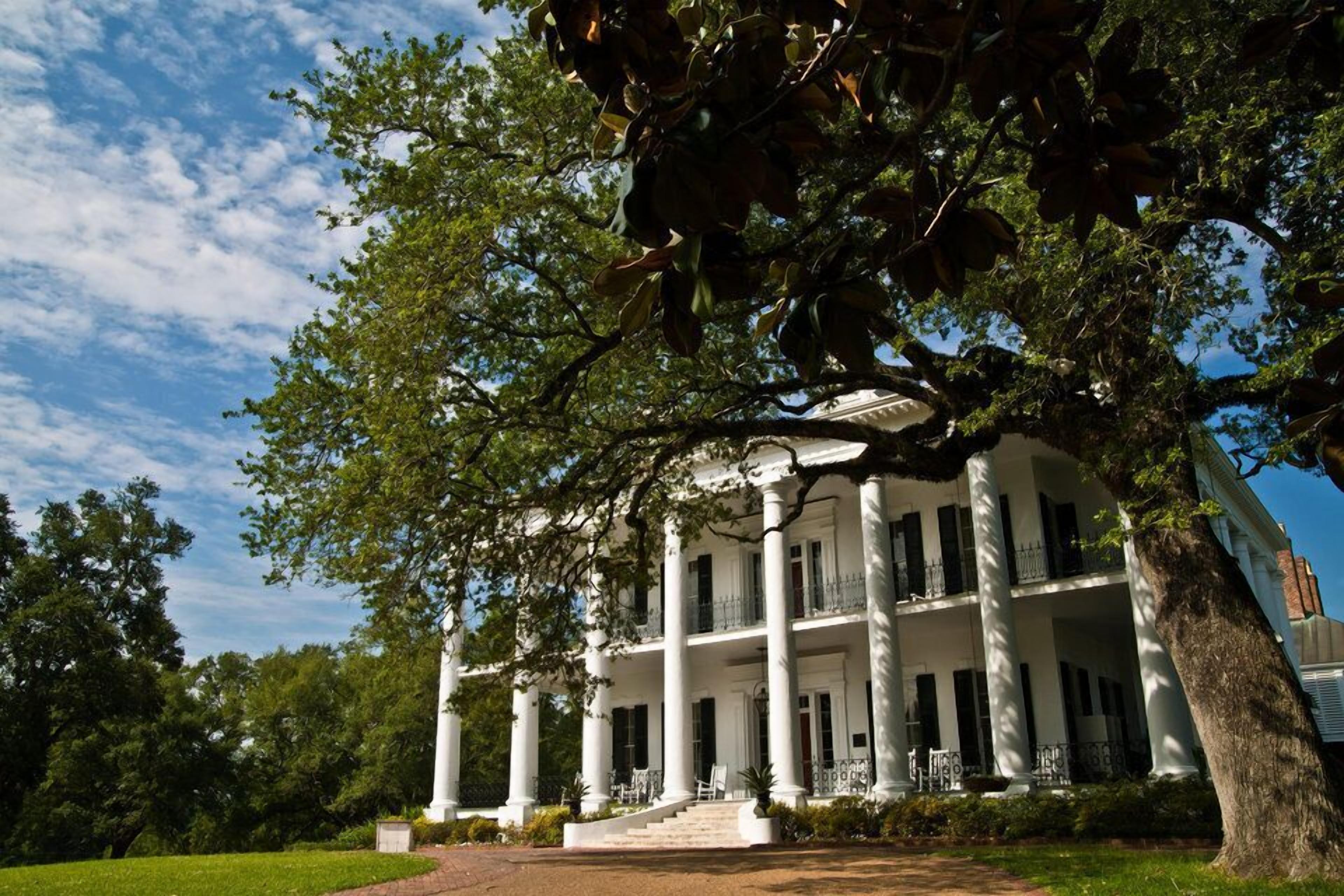 A large, white building with multiple columns and balconies, surrounded by lush greenery and trees.