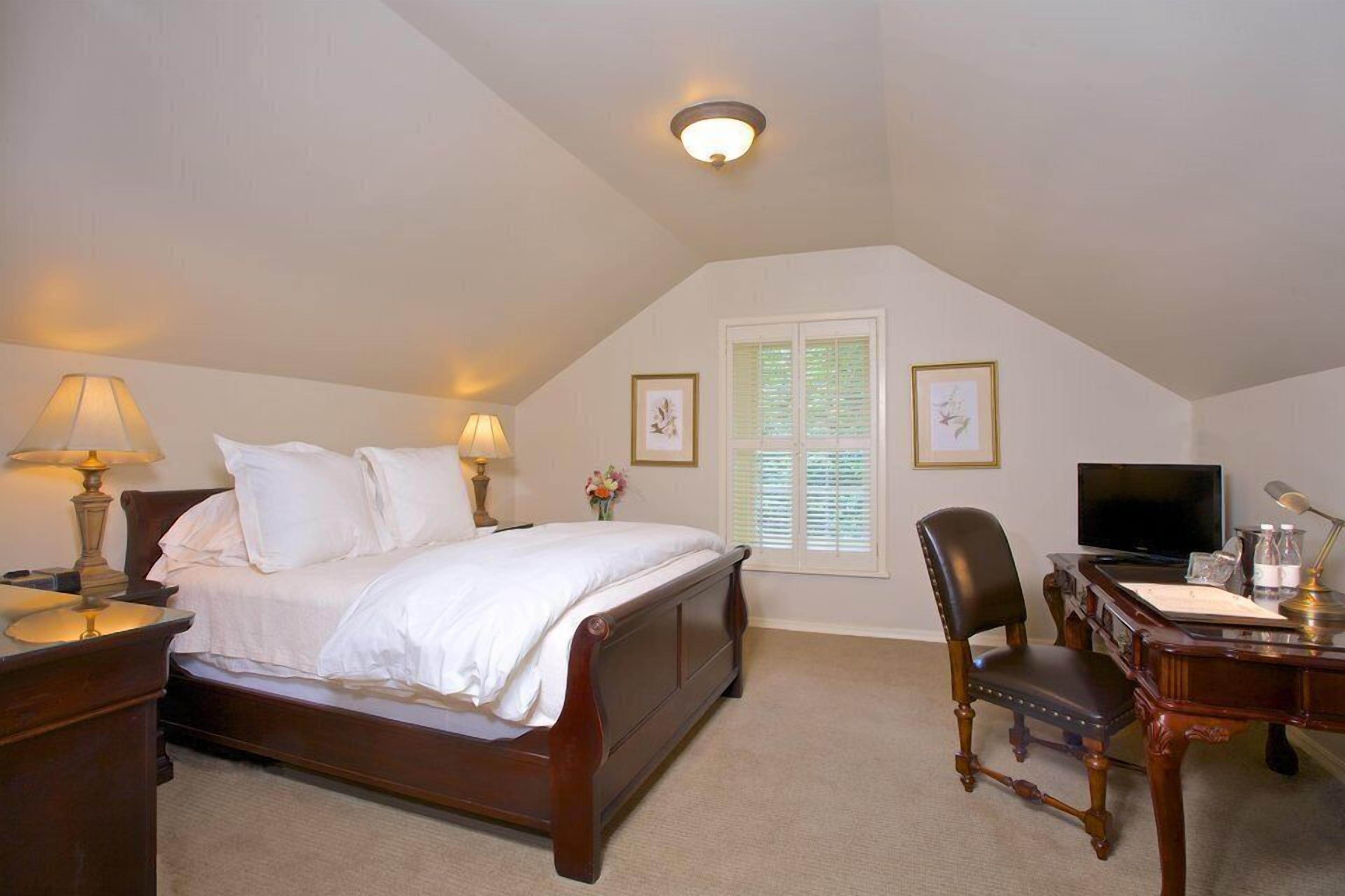 A bedroom with a bed, bedside lamps, a desk with a chair, a television, and framed pictures on the wall.