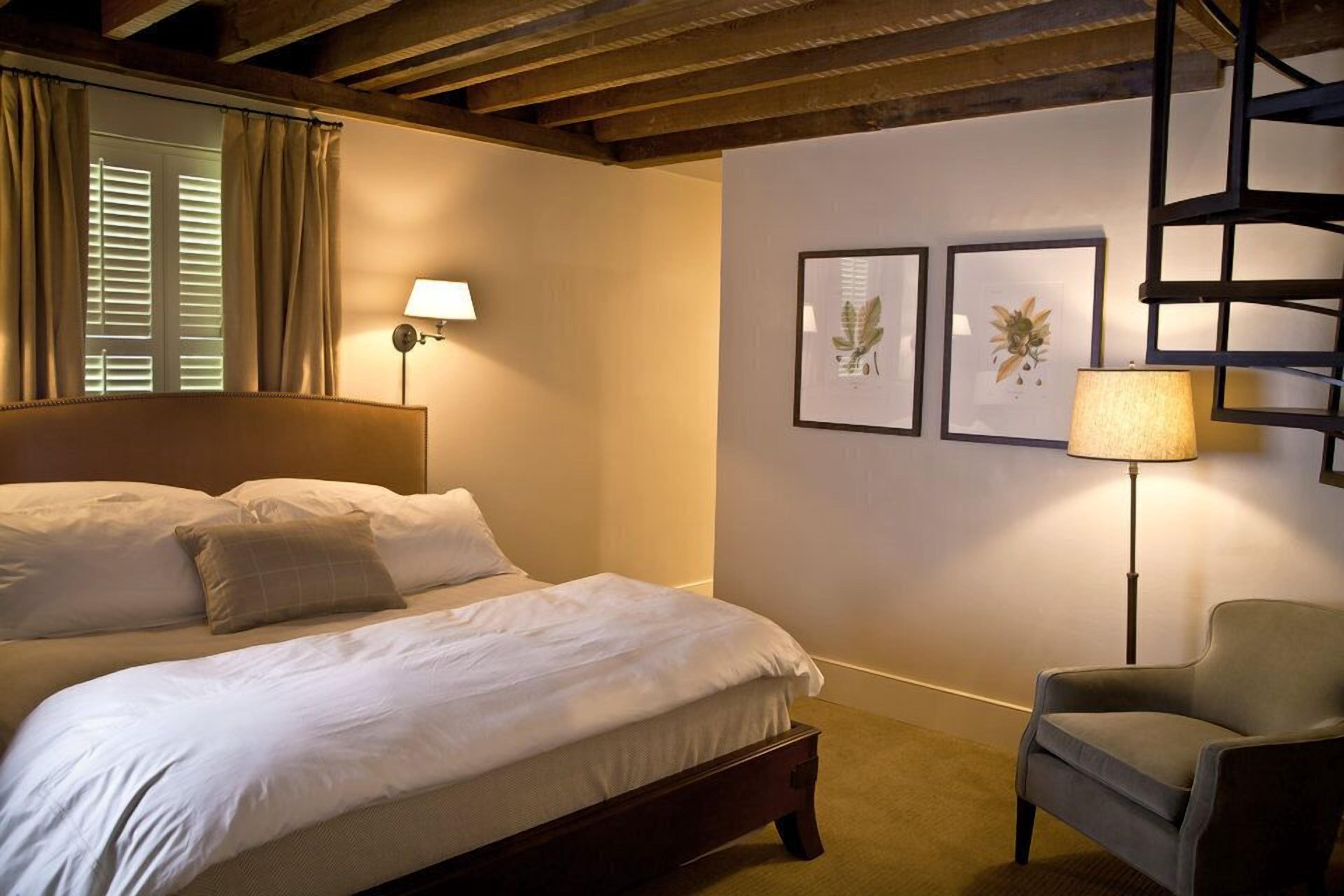 A bedroom with a large bed, a chair, a lamp, and framed botanical prints on the wall.