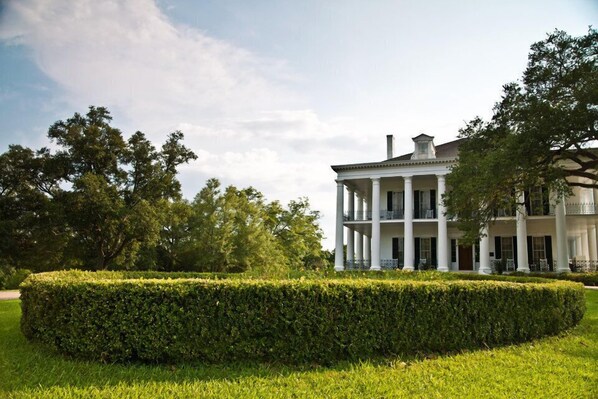 A large, white building with columns and a balcony, surrounded by well-manicured hedges and trees.
