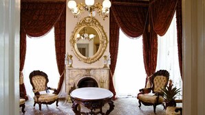 Une pièce avec une cheminée, des fauteuils richement décorés, une table ronde et un grand miroir.