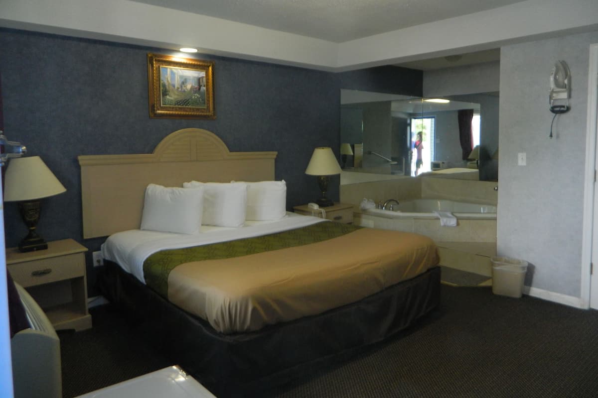 A hotel room with a bed, bedside lamps, a large mirror, a bathroom with a bathtub, and a trash bin.