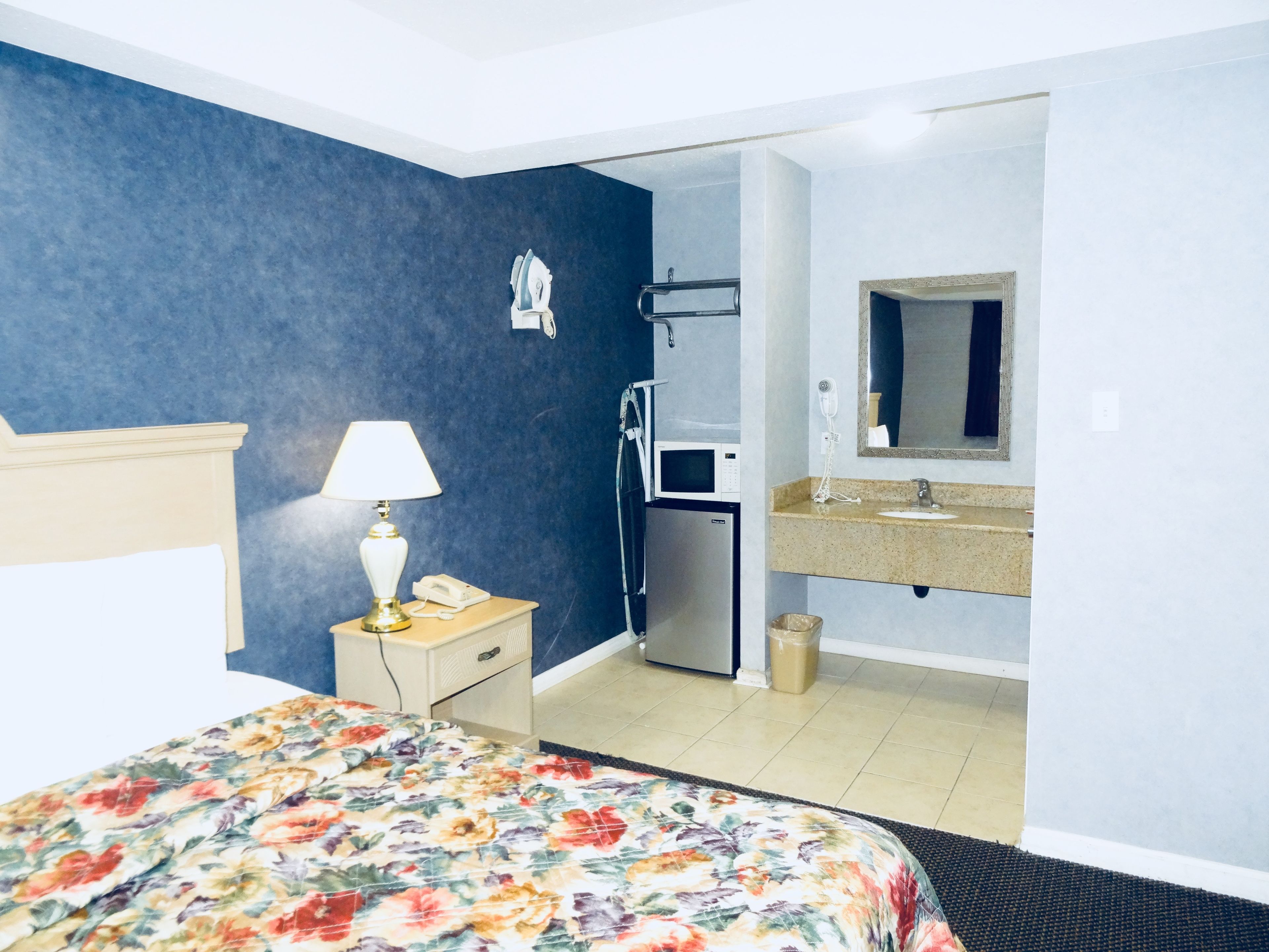 A hotel room with a bed, a bedside lamp, a microwave, a hairdryer, a sink, and a mirror.
