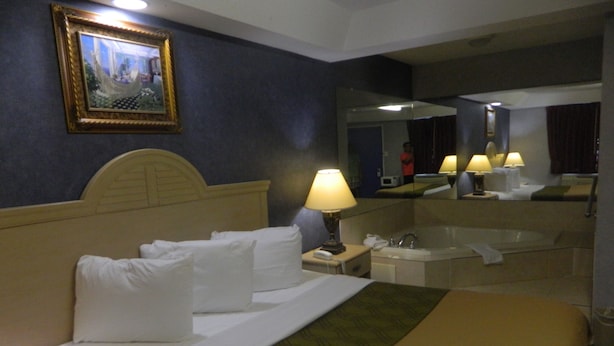 A hotel room with a bed, a nightstand with a lamp, a bathroom with a bathtub, and a painting on the wall. - Village Inn and Suites (Absecon)