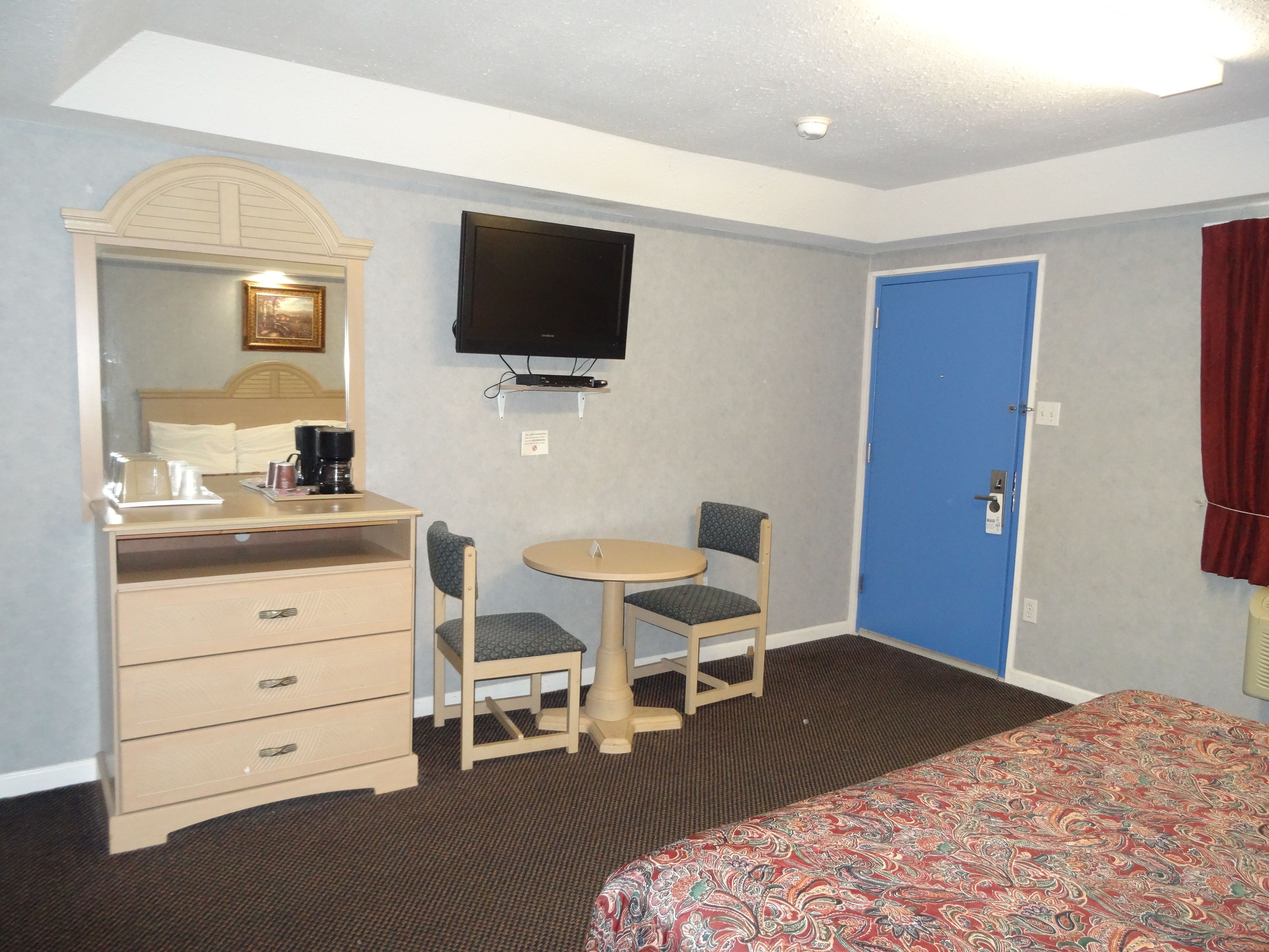 A hotel room with a bed, a desk with a chair, a television, and a blue door.