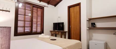Habitación de hotel con cama, ventilador de techo, refrigerador pequeño, puerta de madera y ventana con persianas.