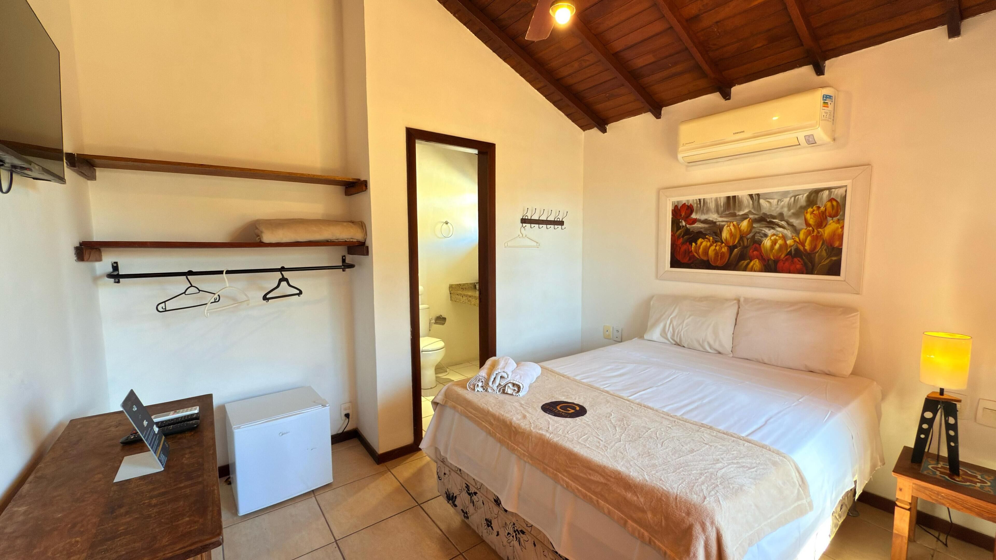 A hotel room with a bed, a small desk with a laptop, a mini-fridge, a shower, and a painting on the wall.