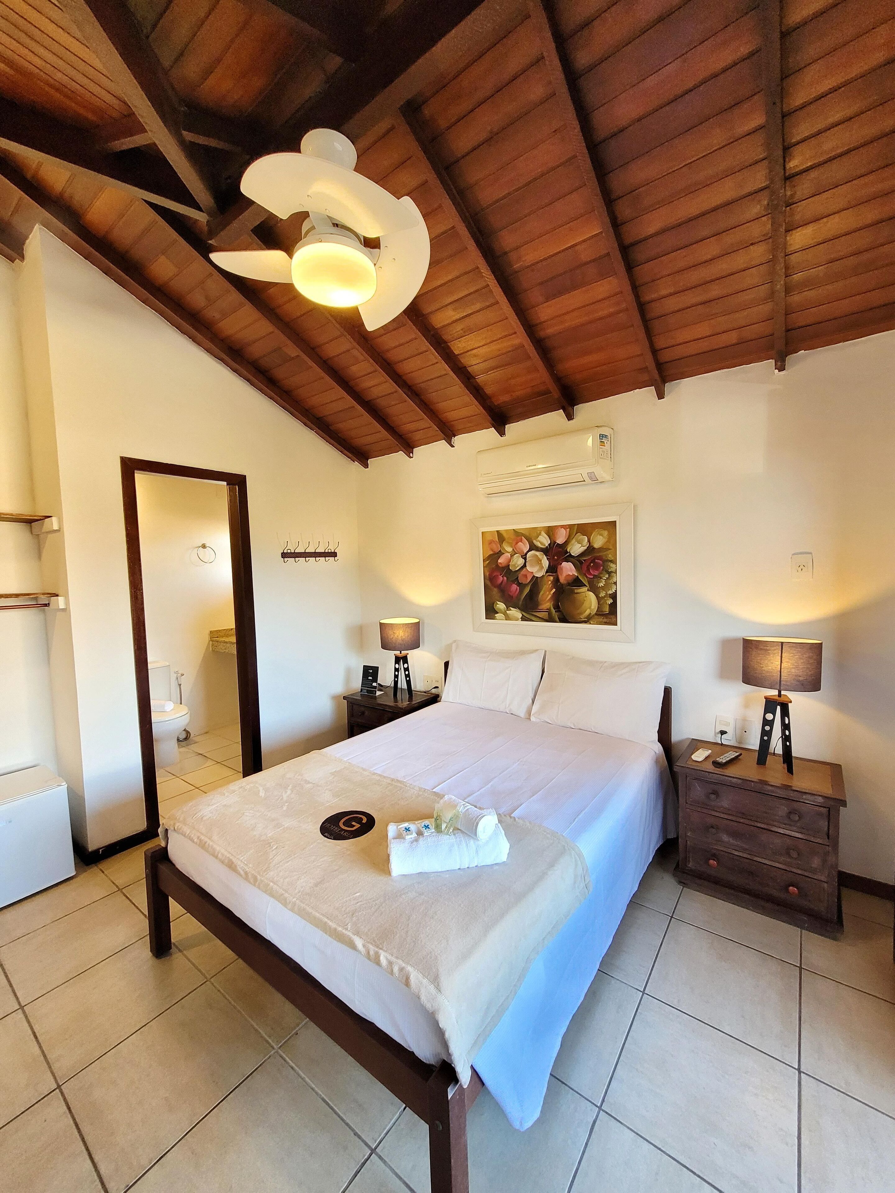 Une chambre d’hôtel avec un plafond en bois, un grand lit recouvert d’une courtepointe blanche, deux tables de chevet avec des lampes, un climatiseur fixé au mur et un tableau au-dessus du lit.