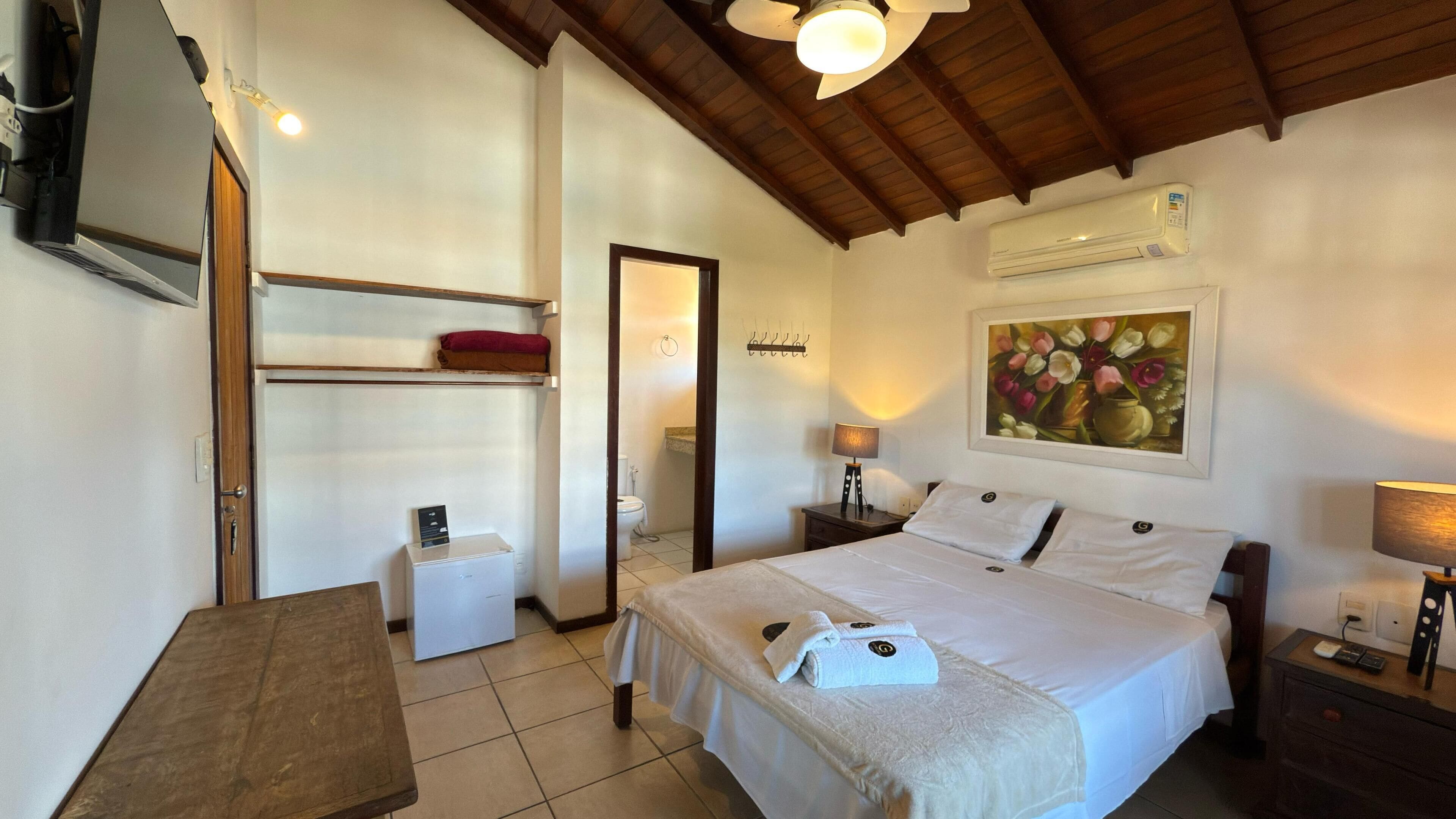 A hotel room with a bed, bedside tables, a desk, a chair, a painting, and an air conditioning unit.
