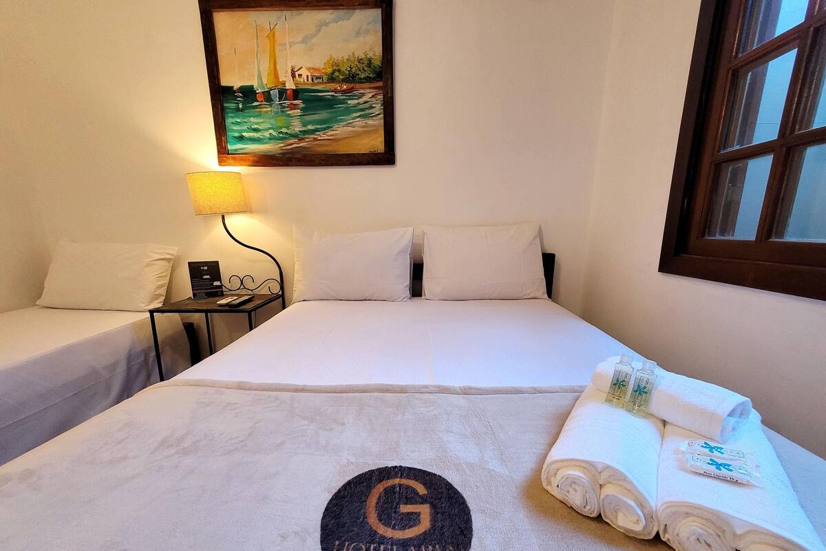 Une chambre d’hôtel avec un lit, une table de chevet, une lampe, un tableau représentant un voilier et une fenêtre.