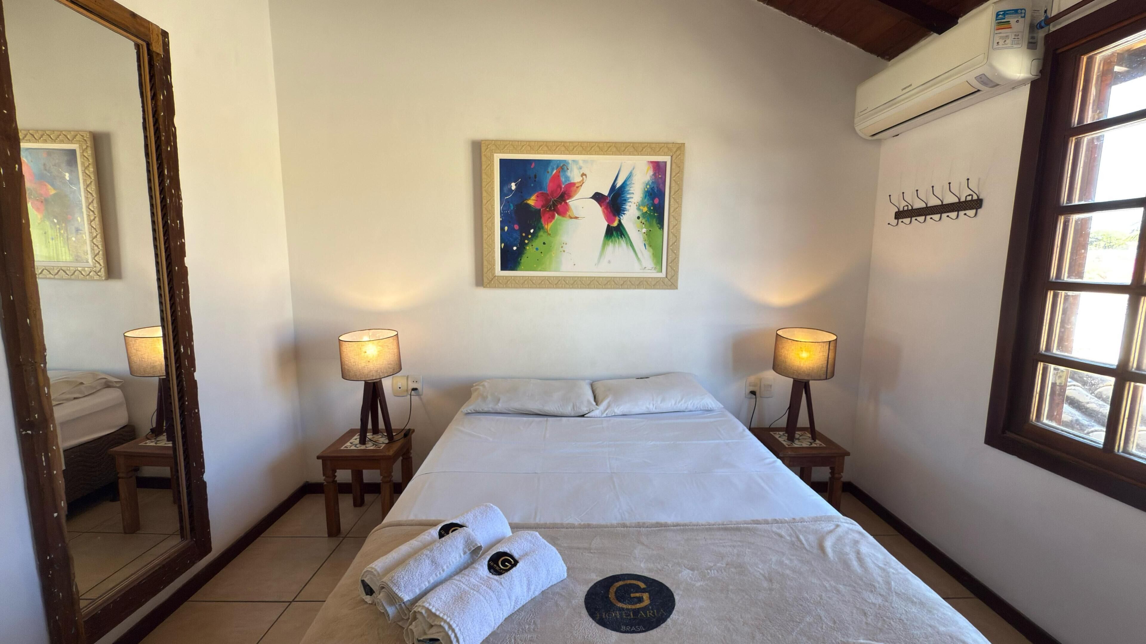 A hotel room with a bed, two bedside lamps, a painting, a mirror, and a window.