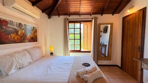 Een slaapkamer met een groot bed, een bloemen schilderij, een raam met jaloezieën, een spiegel en een houten deur.