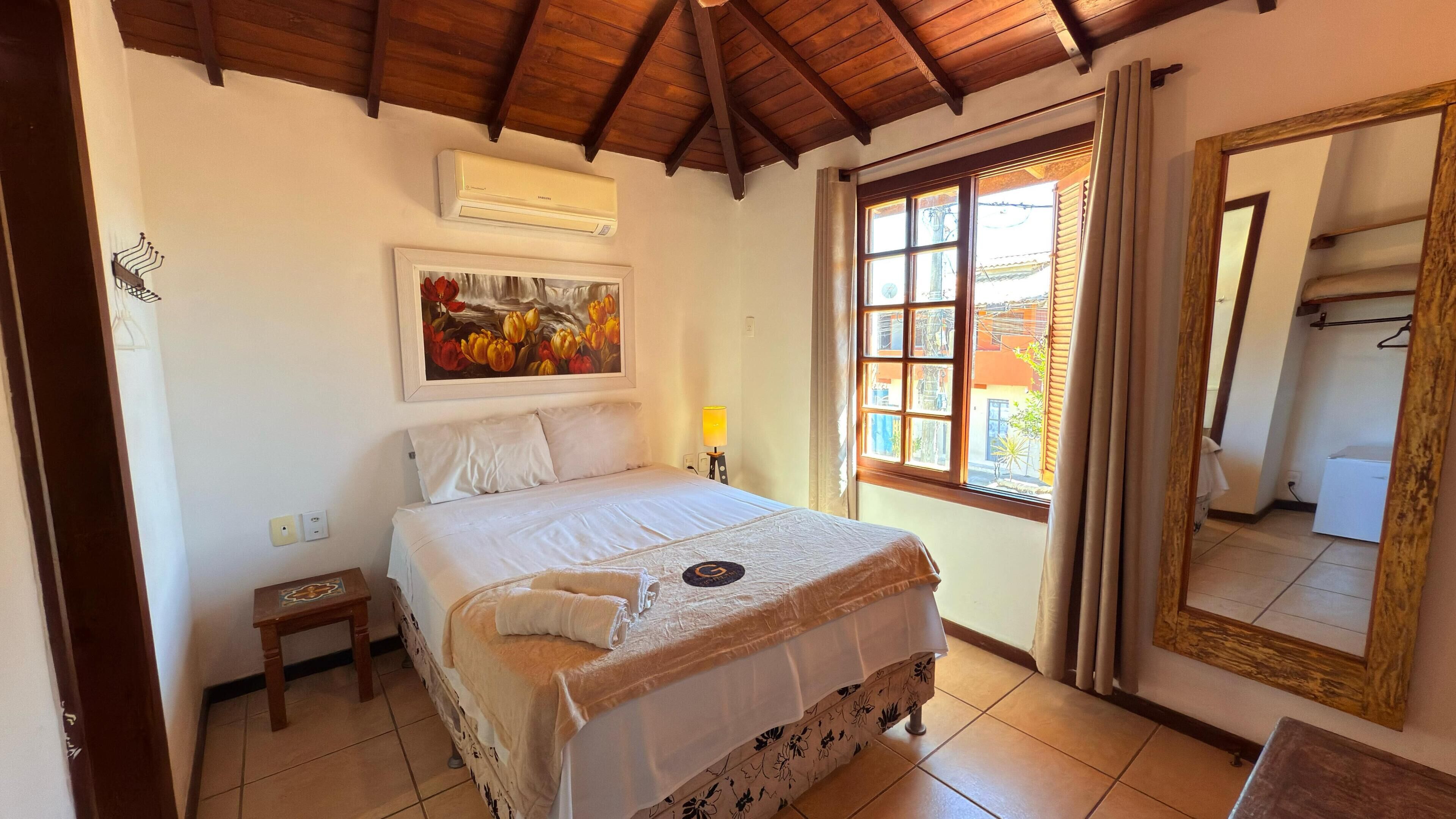 A bedroom with a bed, a nightstand, a lamp, a window with curtains, a painting, and a mirror.