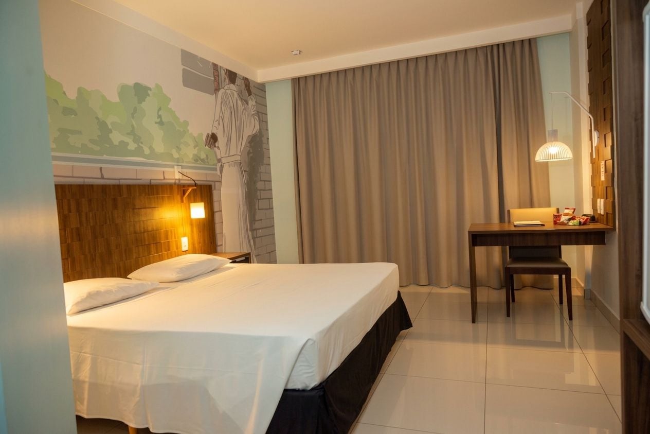 Habitación de hotel con cama, escritorio, silla, lámpara y un amplio ventanal con cortinas.
