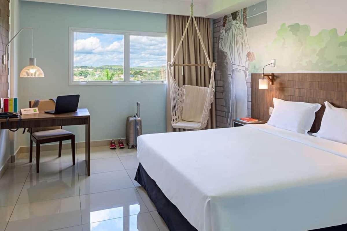 A hotel room with a bed, a desk with a laptop, a hanging chair, a suitcase, and a view of the outdoors.