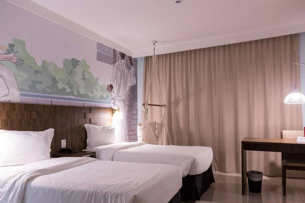 A hotel room with two beds, a wooden headboard, a mural on the wall, a desk, and a hanging chair.