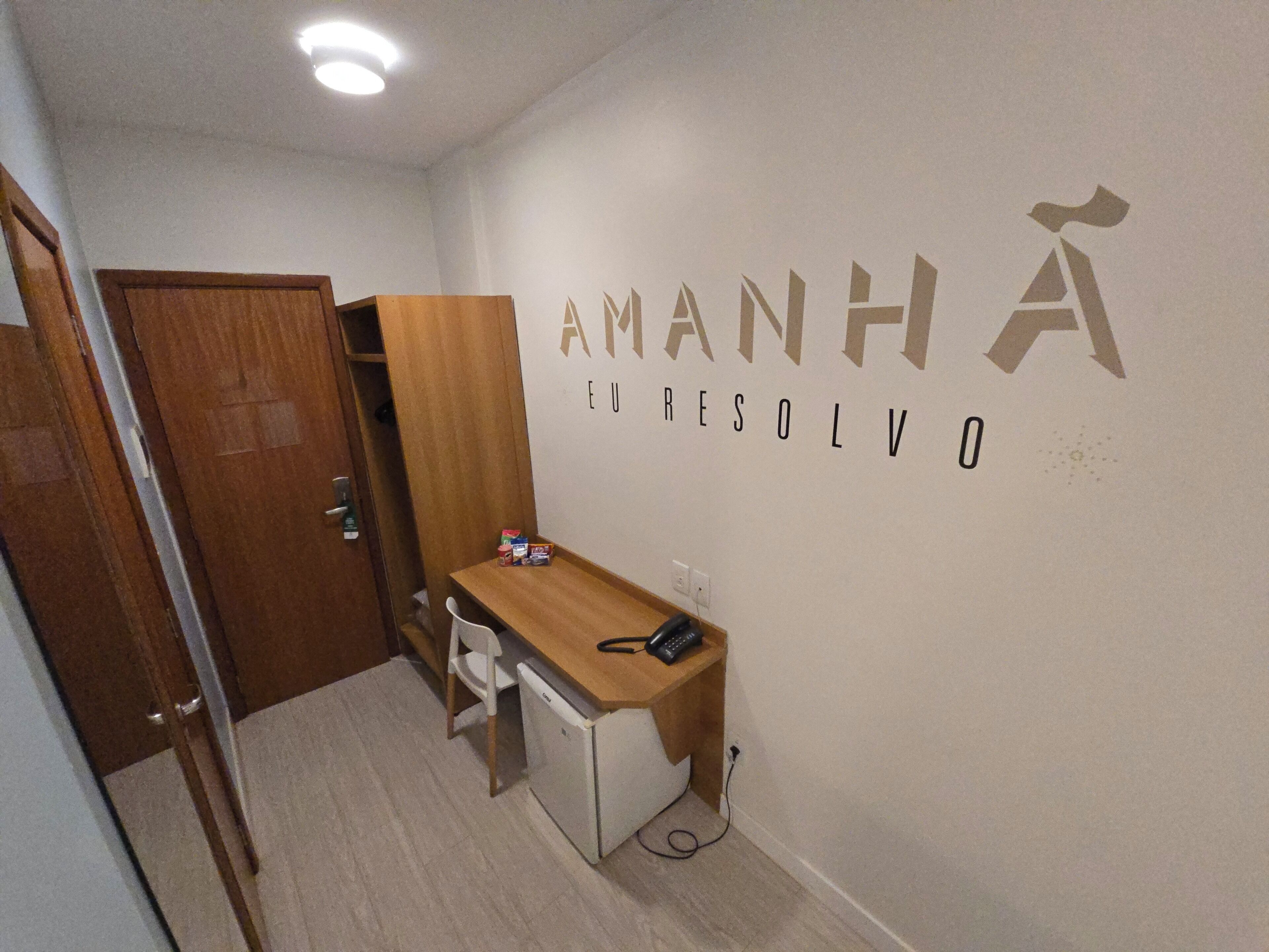A small room with a desk, chair, and a wardrobe. The wall has the text "AMANHA EL RESOLVO" written on it.